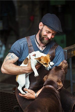 pic of dog and men - A man in a leather apron playing with two dogs in a workshop. Photographie de stock - Premium Libres de Droits, Code: 6118-08729051