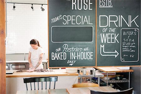 Woman wearing a white apron standing at a work counter in a bakery, menu on a blackboard. Stock Photo - Premium Royalty-Free, Code: 6118-08729119