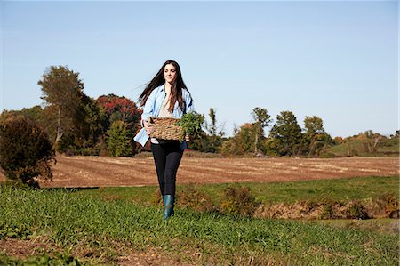 A young woman in working clothes walking across a field, holding a basket of crops. Stock Photo - Premium Royalty-Free, Code: 6118-08842024