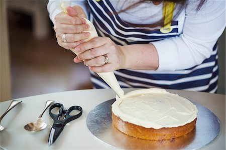 Close up of person wearing a blue and white stripy apron, holding a piping bag, decorating a cake with cream, spoons and scissors on table. Foto de stock - Sin royalties Premium, Código: 6118-08971502