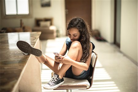 A girl sitting looking at a mobile phone screen with feet up on a kitchen counter. Stock Photo - Premium Royalty-Free, Code: 6118-08991449