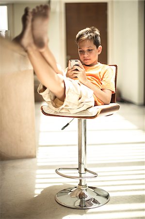 A boy sitting looking at a mobile phone screen with feet up on a kitchen counter. Stock Photo - Premium Royalty-Free, Code: 6118-08991457