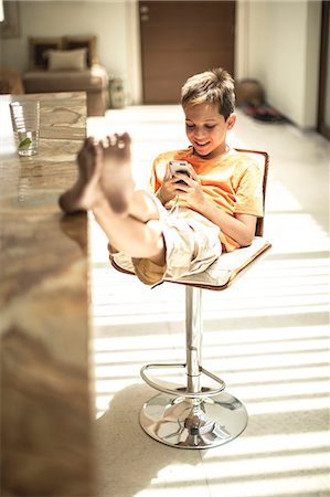 A boy sitting looking at a mobile phone screen with feet up on a kitchen counter. Stock Photo - Premium Royalty-Free, Code: 6118-08991456