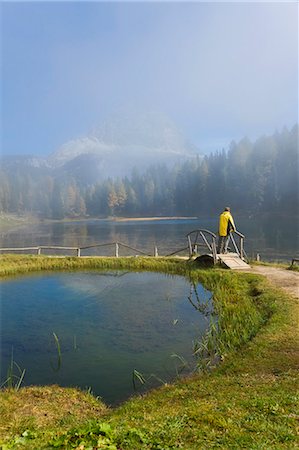 simsearch:6118-08521813,k - Rear view of man standing on wooden bridge over lake, forest and mountain in the distance. Stock Photo - Premium Royalty-Free, Code: 6118-09076452