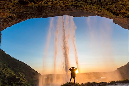 Rear view of man standing underneath natural arch behind waterfall at sunset. Foto de stock - Sin royalties Premium, Código: 6118-09076540