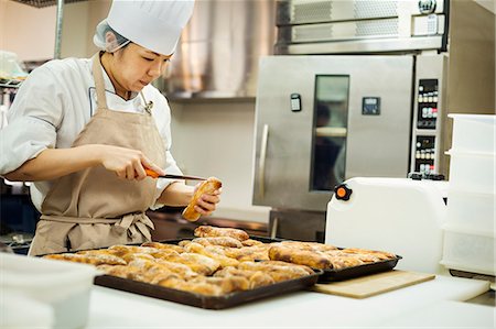 supermarket baker - Woman wearing chef's hat and apron working in a bakery, slicing freshly baked rolls on large trays. Stock Photo - Premium Royalty-Free, Code: 6118-09079247