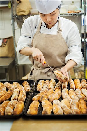 supermarket baker - Woman wearing chef's hat and apron working in a bakery, slicing freshly baked rolls on large trays. Stock Photo - Premium Royalty-Free, Code: 6118-09079246