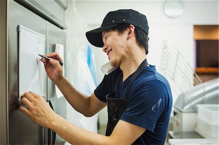 supermarket baker - Man working in a bakery, wearing baseball cap and apron, writing note on small whiteboard, using phone and smiling. Stock Photo - Premium Royalty-Free, Code: 6118-09079251