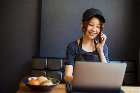 simsearch:614-02985267,k - Woman working in a bakery, wearing baseball cap, sitting at table in front of laptop, using mobile phone. Stock Photo - Premium Royalty-Free, Code: 6118-09079259