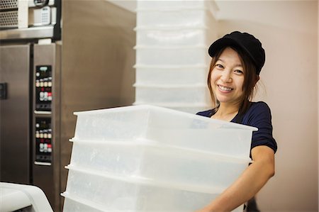 supermarket baker - Woman working in a bakery, wearing baseball cap, carrying stack of white plastic crates. Stock Photo - Premium Royalty-Free, Code: 6118-09079263