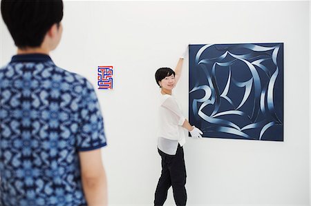 simsearch:693-05552760,k - Rear view of man wearing blue shirt watching woman hang modern painting on white wall in art gallery. Stock Photo - Premium Royalty-Free, Code: 6118-09079482