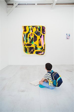 simsearch:693-05552760,k - Boy with short black hair wearing backpack sitting on floor in art gallery with pen and paper, looking at modern painting. Stock Photo - Premium Royalty-Free, Code: 6118-09079487
