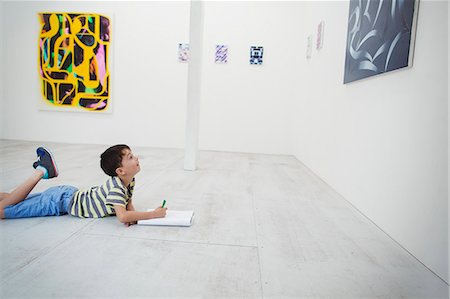 Boy with short black hair lying on floor in art gallery with pen and paper, looking at modern painting. Stock Photo - Premium Royalty-Free, Code: 6118-09079497