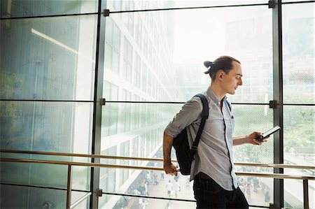 A young Caucasian man with a backpack on a walkway over a road looking at his smart phone, finding his way or texting. Stock Photo - Premium Royalty-Free, Code: 6118-09079725