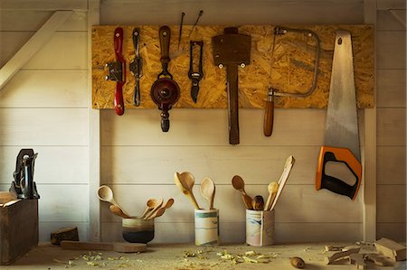 Ceramic pots with a selection of finished handmade wooden spoons and hand tools for carving and cutting wood hanging on a toolboard on a wall above the workbench in a craftsman's workshop. Stock Photo - Premium Royalty-Free, Code: 6118-09018591