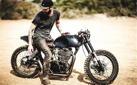 Side view of bearded man with tattoos on his arms sitting on cafe racer motorcycle on a dusty dirt road. Stock Photo - Premium Royalty-Free, Code: 6118-09027929