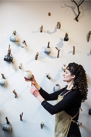 dexterity - Close up of woman with curly brown hair wearing apron standing in pottery shop, hanging mugs onto small branches on a wall. Stock Photo - Premium Royalty-Free, Code: 6118-09148233