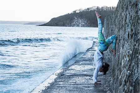 simsearch:6118-07731916,k - Young woman doing handstand on a wall by the ocean, one leg raised. Photographie de stock - Premium Libres de Droits, Code: 6118-09165863