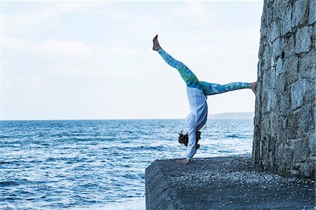 simsearch:6118-07731916,k - Young woman doing handstand on a wall by the ocean, one leg raised. Photographie de stock - Premium Libres de Droits, Code: 6118-09165862