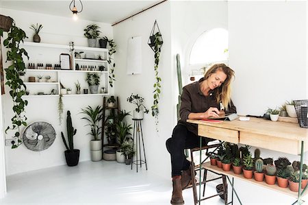 simsearch:6118-08202525,k - Female owner of plant shop sitting at table, working on digital tablet, a selection of plants on wooden shelves. Stock Photo - Premium Royalty-Free, Code: 6118-09165898