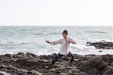 simsearch:6118-07731916,k - Young woman with brown hair and dreadlocks wearing white blouse standing on rocky shore by ocean, doing Tai Chi. Photographie de stock - Premium Libres de Droits, Code: 6118-09165827