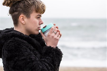 simsearch:400-04435419,k - Portrait of young woman with brown hair and dreadlocks and a lip piercing wearing black furry jacket, standing by ocean, holding mug to her lips. Stock Photo - Premium Royalty-Free, Code: 6118-09165830