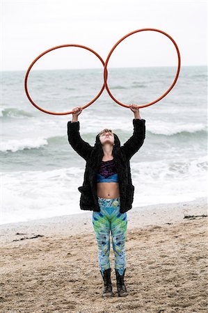 simsearch:6118-07731916,k - Young woman with brown hair and dreadlocks standing on a sandy beach by the ocean, balancing two hula hoops. Photographie de stock - Premium Libres de Droits, Code: 6118-09165832