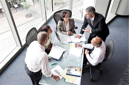 A mixed race group of male and female business people in a meeting at a conference table next to a large window in a convention centre. Stock Photo - Premium Royalty-Free, Code: 6118-09174115
