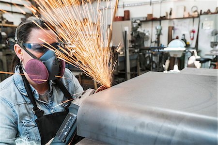 Woman wearing safety glasses and dust mask standing in metal workshop, using power grinder, sparks flying. Stock Photo - Premium Royalty-Free, Code: 6118-09112050