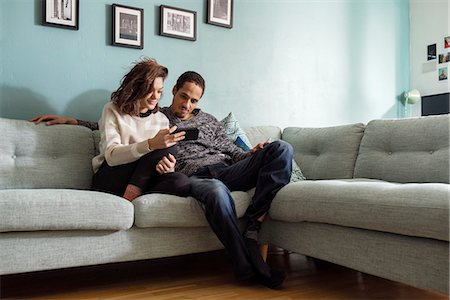Sweden, Young couple sitting on sofa and looking at smart phone Stock Photo - Premium Royalty-Free, Code: 6126-08659313