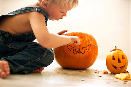 Young boy kneeling on the floor leans forward to carve the face of a Jack O'Lantern with a tool on a big pumpkin with a finished small Jack O'Lantern next to him. Stock Photo - Premium Royalty-Free, Code: 6128-08747701