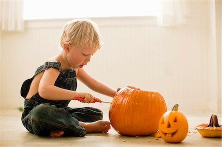 Young boy sitting cross-legged on the floor leans forward to carve the face of a Jack O'Lantern with a knife on a big pumpkin with a finished small Jack O'Lantern next to him. Stock Photo - Premium Royalty-Free, Code: 6128-08747702