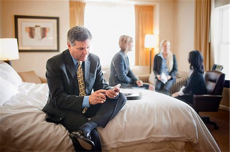 Businessman sits on the edge of a bed in a hotel room during a business trip and texts on his cell phone while his three female colleagues work together in the background. Stock Photo - Premium Royalty-Free, Code: 6128-08766783