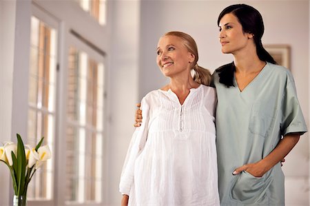 Female nurse standing with her arm around senior female patient as they look out window. Photographie de stock - Premium Libres de Droits, Code: 6128-08780789