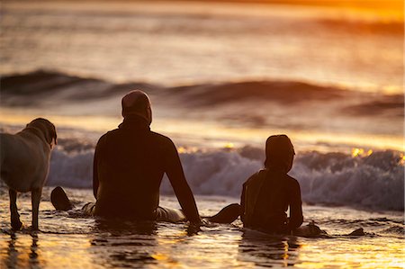 rear of man sitting - Mid adult man and his young son sitting in shallow water on a beach with their dog. Stock Photo - Premium Royalty-Free, Code: 6128-08738251