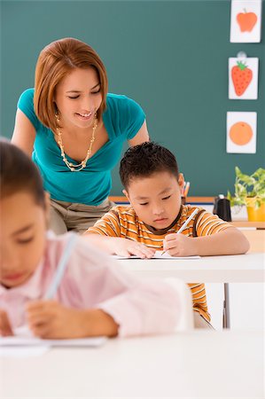 Close-up of a schoolboy writing on a notebook with a pencil with his female teacher standing behind him Stock Photo - Premium Royalty-Free, Code: 625-02930457