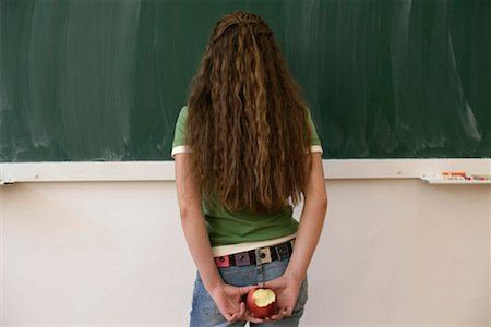 Teenage girl standing in front of a blackboard, holding an apple behind her back Stock Photo - Premium Royalty-Free, Code: 628-00920631