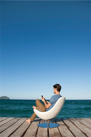 Young man sitting in armchair by lake using cell phone, side view Stock Photo - Premium Royalty-Free, Code: 632-05817150
