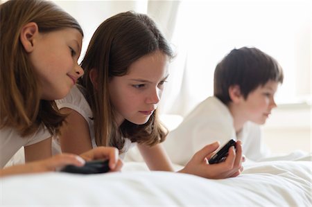 simsearch:632-03424261,k - Sisters lying on bed playing handheld video game, boy in background Stock Photo - Premium Royalty-Free, Code: 632-06029927