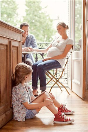 daughter sitting on dad lap - Girl sitting on floor with digital tablet as parents chat in background Stock Photo - Premium Royalty-Free, Code: 632-08545922