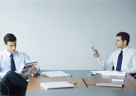 Business colleagues sitting at table, one reading magazine while other checks messages on cell phone Stock Photo - Premium Royalty-Free, Code: 632-01155403