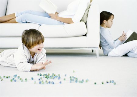 Boy playing marbles on floor, woman and girl reading in background Stock Photo - Premium Royalty-Free, Code: 632-01151560