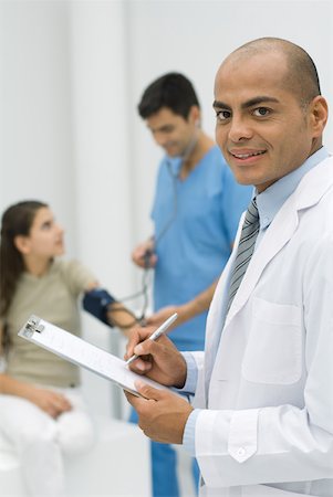 simsearch:694-03331631,k - Doctor writing on clipboard, smiling at camera, nurse measuring patient's blood pressure in background Stock Photo - Premium Royalty-Free, Code: 632-02227628