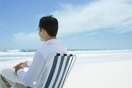 Man sitting in folding chair on beach, using pen and paper, rear view Stock Photo - Premium Royalty-Free, Code: 633-01713847