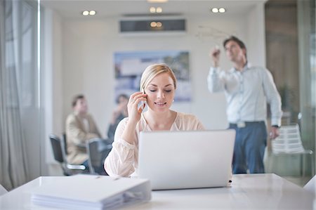 simsearch:649-07238396,k - Woman sitting at conference room table using laptop and mobile phone, colleagues working in background Stock Photo - Premium Royalty-Free, Code: 649-07238398