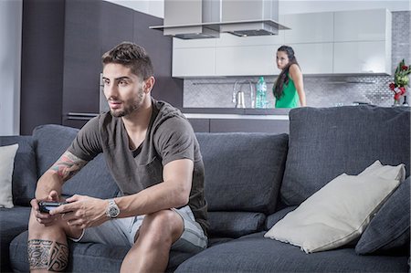 Young man on sofa using gaming control watched by angry girlfriend Stock Photo - Premium Royalty-Free, Code: 649-08179946
