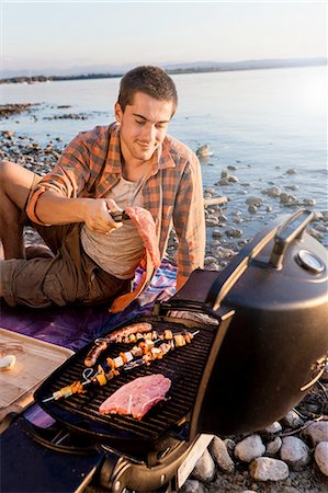 Young man sitting next to water cooking meat and kebabs on barbecue, Schondorf, Ammersee, Bavaria, Germany Stock Photo - Premium Royalty-Free, Code: 649-08307282