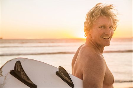 Man on beach carrying surfboard looking over shoulder at camera smiling Stock Photo - Premium Royalty-Free, Code: 649-08543785