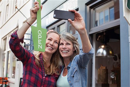 simsearch:614-07806416,k - Women in front of shop holding open sign taking selfie with smartphone Photographie de stock - Premium Libres de Droits, Code: 649-08576889