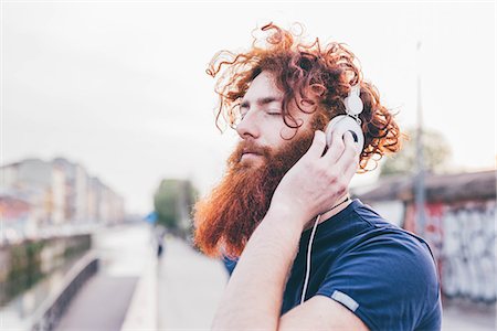 Young male hipster with red hair and beard listening to headphones with eyes closed in city Stock Photo - Premium Royalty-Free, Code: 649-08702677